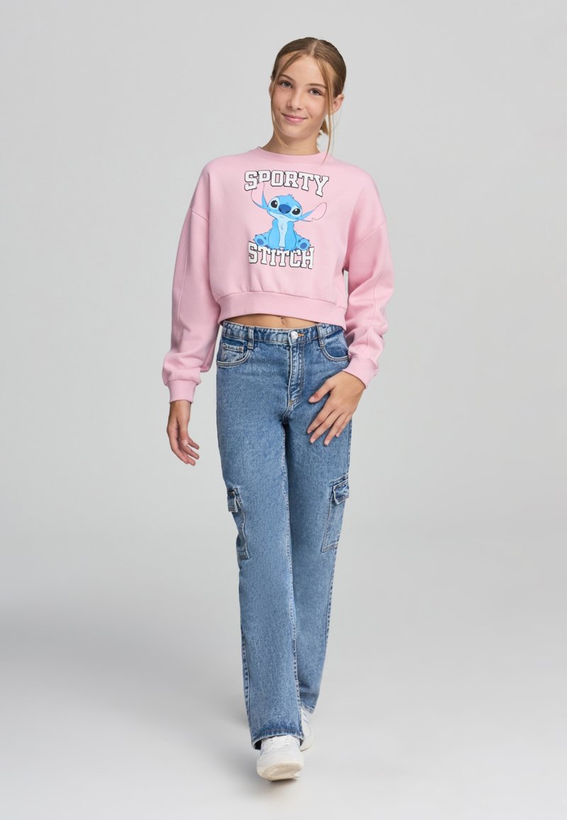 Różowy krótki sweatshirt z niebieską postacią z kreskówki oraz napisem "SPORTY STITCH". Połączony z jasnoniebieskimi luźnymi dżinsami i białymi trampkami.