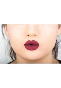 Rouge à lèvres mat dans une teinte bordeaux profond appliqué sur des lèvres pleines et bien définies. Texture lisse avec un fini velouté, accentuant les contours des lèvres.