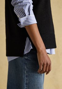 Arm und Hand einer Person, die ein blau-weiß gestreiftes Hemd mit hochgekrempelten Ärmeln, einen schwarzen ärmellosen Pullover und blaue Jeans trägt.