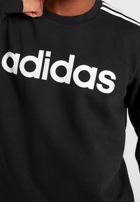 Sweat-shirt noir avec logo "adidas" blanc, col rond et trois bandes blanches sur l'épaule. Le tissu semble doux et lisse.