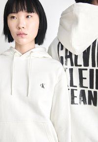 Felpa bianca con cappuccio, dotata di tasca frontale a marsupio e cordoncino per regolare il cappuccio. Sul retro presenta lettering nero in grassetto.