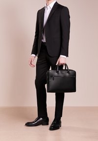 Mand i en sort jakkesæt og hvid skjorte holder en sort læder portefølje, står på en beige gulv mod en bleg baggrund.