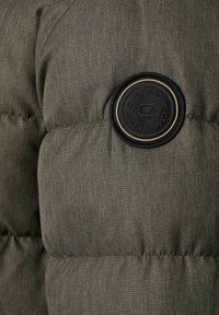 Quiltet olivengrøn dunjakkeafsnit med en tekstureret finish, der har en rund sort logo-patch på ærmet.