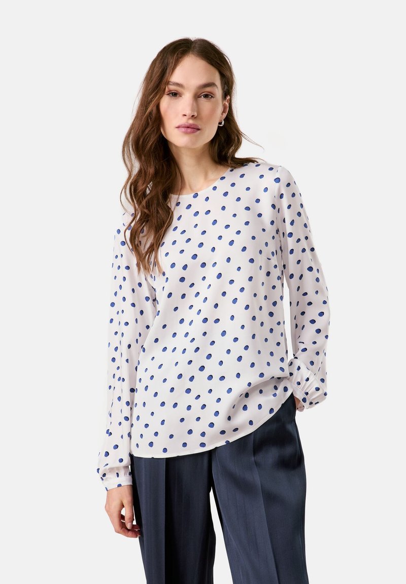 White long-sleeve blouse with blue polka dots, round neck, and gathered sleeves, paired with navy trousers. Smooth, lightweight fabric.