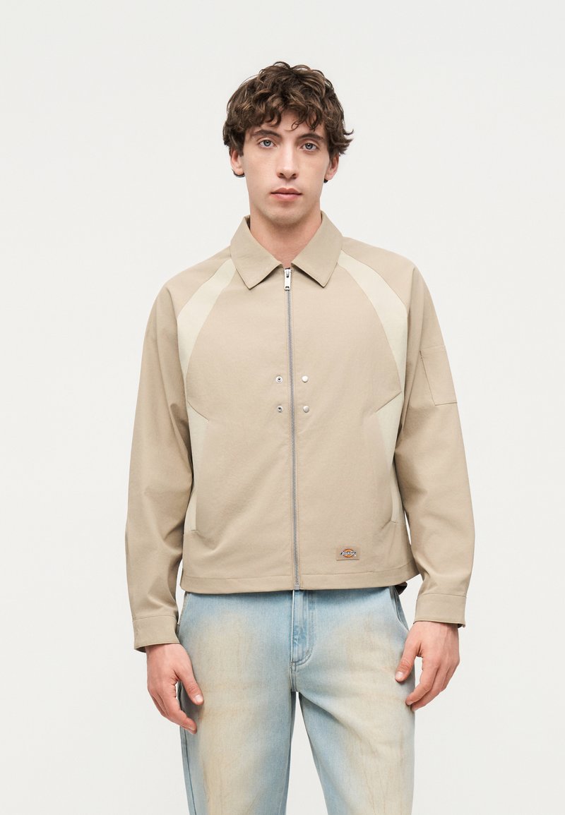 Jeune homme debout, portant une veste beige zippée avec des détails de boutons et un jean bleu clair délavé sur fond uni.