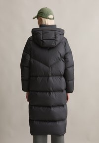 Lang zwarte dik gewatteerde jas met een capuchon, voorzien van een gequilte uitstraling en zichtbare naden. Draagt over grijs geruite broek. Pet zichtbaar op het hoofd.