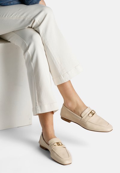 Mocassins en cuir beige avec un design à orteils carrés, présentant un détail en métal doré et un petit talon en bois. Portés avec un pantalon blanc.