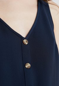 Mörkblå ärmlös topp med V-ringning, texturerad tyg och två guldtonade knappar längs framstycket.