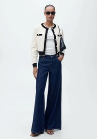 Cardigan crème et noir avec des boutons dorés, haut blanc, pantalon en denim foncé à jambes larges et sac à main noir. Sandales plates à bout fermé.