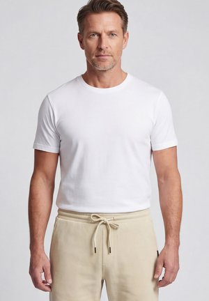 Homme adulte aux cheveux courts bruns, portant un t-shirt blanc uni et un pantalon beige avec cordon, debout devant un fond gris neutre.