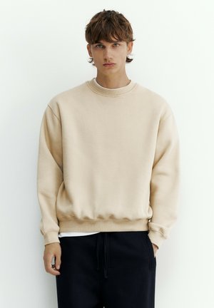 Junge Person mit kurzen braunen Haaren, die einen beigen Pullover und schwarze Hose trägt, steht vor einem schlichten weißen Hintergrund.