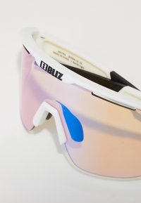 Sportsonnenbrille mit großem, gebogenem, pink getöntem Glas, weißem Rahmen und schwarzen Akzenten. Verfügt über gummierte Nasenpads für besseren Halt. Logo oben angebracht.