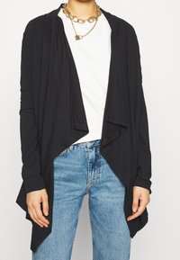 Persona che indossa un cardigan nero aperto nella parte anteriore, una camicia bianca, jeans blu e una collana d'oro massiccia con un piccolo ciondolo a forma di lucchetto.