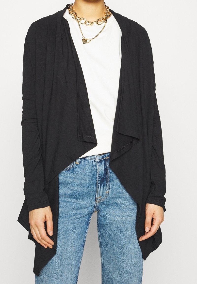 Persona che indossa un cardigan nero aperto nella parte anteriore, una camicia bianca, jeans blu e una collana d'oro massiccia con un piccolo ciondolo a forma di lucchetto.