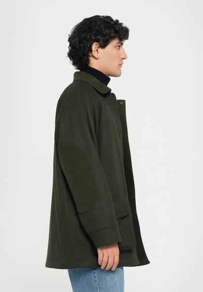 Olive green long coat with a collared neckline, snap buttons, and textured fabric. Features suede-like elbow patches and front pockets.