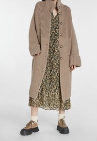 Cardigan long en tricot beige épais avec un col haut, de grands boutons et des poignets retournés, porté sur une robe fleurie et associé à des chaussures brunes.