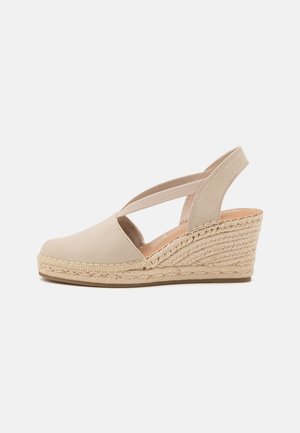 Espadrille à talon compensé beige avec bout fermé, sangle élastique croisée et semelle en jute tressé sur fond blanc.