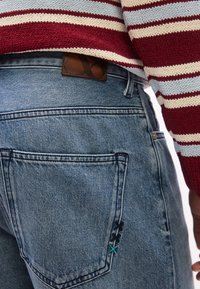 Πίσω μέρος ατόμου που φοράει μπλε τζιν με κεντημένη τσέπη και πουλόβερ πλεκτό με ρίγες κόκκινες, λευκές και γαλάζιες.