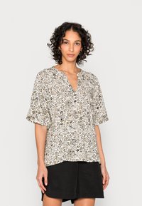 Blusa de manga curta, em creme, com um padrão floral em bege e verde, apresentando um decote em V e um corte descontraído, combinada com calções pretos.