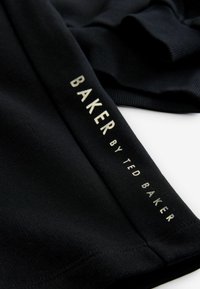 Sort stof med ribbet tekstur, der har teksten "BAKER by TED BAKER" i metallisk guld. Enkel, minimalistisk design, foldet.