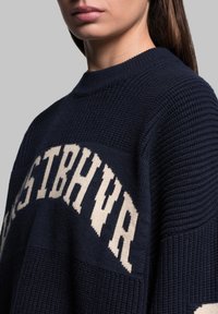 Maglione a maglia navy con colletto rotondo a coste e testo grafico a contrasto in crema. Motivo testurizzato con sezioni a blocchi e design a vestibilità morbida.