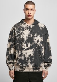 Tânăr îmbrăcat cu un hanorac tie-dye negru și bej, cu buzunar frontal, stând în fața unui fundal gri deschis simplu.