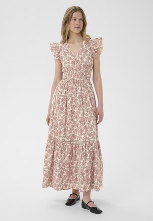 Femme portant une robe maxi blanche sans manches avec des motifs floraux rouges et des épaules volantées, associée à des chaussures plates noires, debout devant un fond uni.