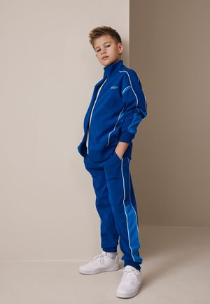 Chlapec stojaci pri béžovej stene, oblečený v modrej teplákovej súprave s bielymi lemami a bielymi teniskami, ruky v vreckách, pozerá pred seba.