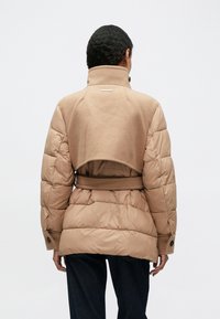 Chaqueta de plumas beige con un alto cuello, cintura ceñida y una textura suave y acolchada. Presenta botones oscuros y una parte superior de lana recortada.