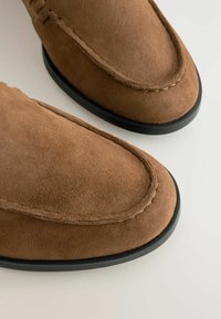 Des chaussures mocassins en daim marron avec un bout arrondi, une semelle en caoutchouc noir et des détails de couture visibles le long des bords. Texture lisse sur l'ensemble.