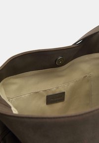 Smeđa platnena torba s mekom teksturom, s bež podstavom i etiketom unutra. Ima kopču na pritisak za siguran pristup.