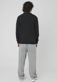 Personne avec des cheveux bouclés portant un pull noir, un jean gris ample et des baskets blanches, se tenant de face devant un fond gris clair uni.