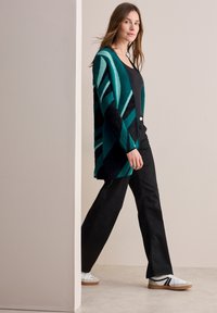 Flerfärgad cardigan med ränder i teal och svart, tillverkad av mjukt material. I kombination med en svart topp och vida mörka byxor.