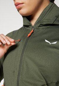 Mørkegrønn hettegenser med teksturert overflate, med en oransje glidelåsdrager og en hvit logo. Nærbilde av glidelåsområdet og hetten.