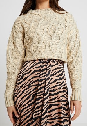 Kobieta ma na sobie kremowy sweter z warkoczowym splotem, zestawiony z beżowo-czarną spódnicą w zebra print, związaną w talii pasem z dopasowanego materiału.