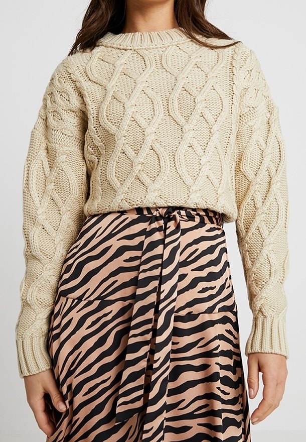 Une femme porte un pull en maille torsadée crème assorti à une jupe imprimée zèbre beige et noire, nouée à la taille avec une ceinture en tissu assortie.