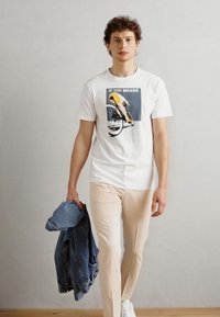 Hvit bomulls t-skjorte med et grafisk motiv av en syklist og teksten "HVIS DU BREKER", kombinert med lysebeige bukser og en denimjakke.