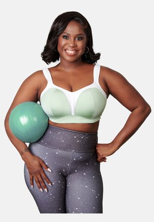 Lächende Frau in grünem Sport-BH und gemusterten Leggings hält einen grünen Gymnastikball unter dem Arm vor einem einfarbigen Hintergrund.