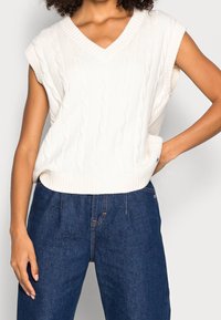 Weißer gestrickter ärmelloser Pullover mit V-Ausschnitt, der ein Zopfmuster und einen gerippten Saum aufweist. Kombiniert mit hochtaillierten dunklen Jeans.
