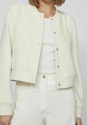 Vrouw draagt een crèmekleurig jasje met textuur en drukknopen over een geribd wit topje en een witte broek met hoge taille, tegen een effen grijze achtergrond.