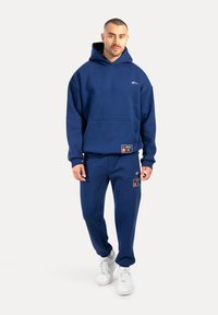 Felpa con cappuccio blu navy con tasca a marsupio, abbinata a pantaloni della tuta coordinati con dettaglio di logo e toppa. Indossata con sneakers bianche.