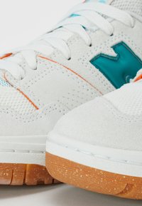 Gros plan sur des baskets blanches en daim avec semelles en gomme, lacets blancs, logo bleu sarcelle et détails de couture orange.