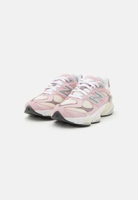 Sapatilhas desportivas rosa e creme com painéis de camurça e malha, apresentando uma sola branca grossa e pormenores cinzentos. Os atacadores são brancos, com um logótipo na lateral.