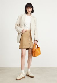 Blazer beige sopra una camicia bianca, gonna a bottoni color sabbia e sneakers bianche. Tiene in mano una borsa arancione. Sfondo con superficie piatta.