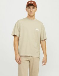 Beige short-sleeve t-shirt with a relaxed fit; features a small white logo. Paired with light beige pants and a brown cap.