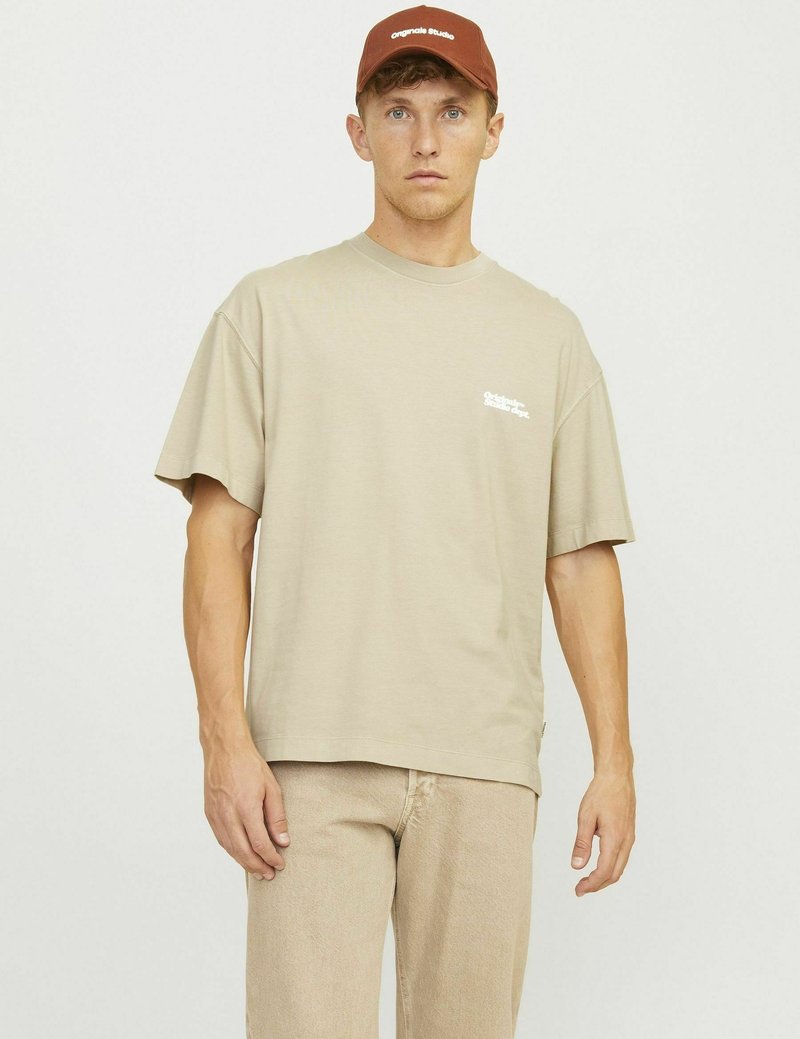Beige short-sleeve t-shirt with a relaxed fit; features a small white logo. Paired with light beige pants and a brown cap.