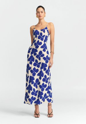 Sukienka maxi ma kremową bazę z dużymi niebieskimi kwiatowymi wzorami, cienkimi ramiączkami i dopasowanym krojem. Gładki materiał i długość do kostek.