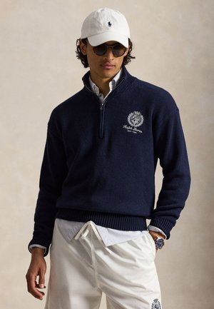 Homme portant une casquette blanche, des lunettes de soleil foncées, un pull marine à fermeture éclair quart de zip avec emblème, une chemise bleu clair, un pantalon blanc à cordon et une montre-bracelet.