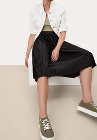 Gonna nera plissè, top verde oliva, giacca di jeans bianca e sneakers verdi oliva con suole bianche. Tessuti lisci, design a strati.