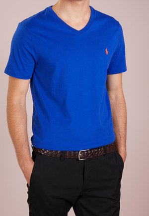 Homme portant un T-shirt bleu vif à col en V avec un petit logo brodé, pantalon foncé, ceinture tressée marron, mains dans les poches.